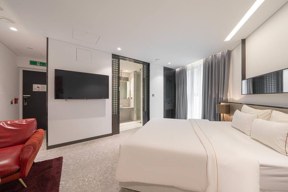 A modern hotel room with a large bed, a flat-screen TV, a red leather chair, and a glass partition door leading to a bathroom.