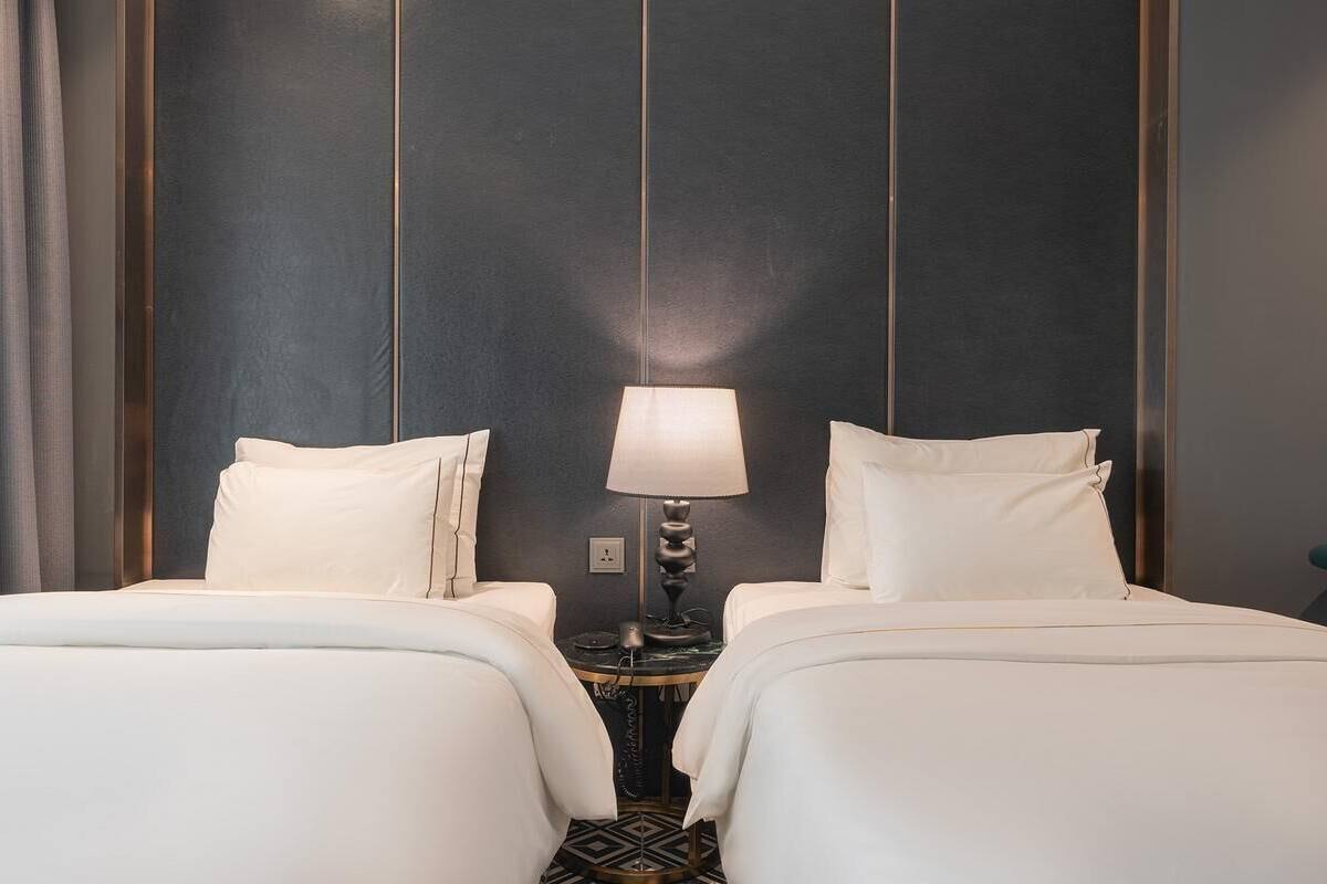 A hotel room with two beds, a round table with a lamp, and a dark wall with vertical lines.