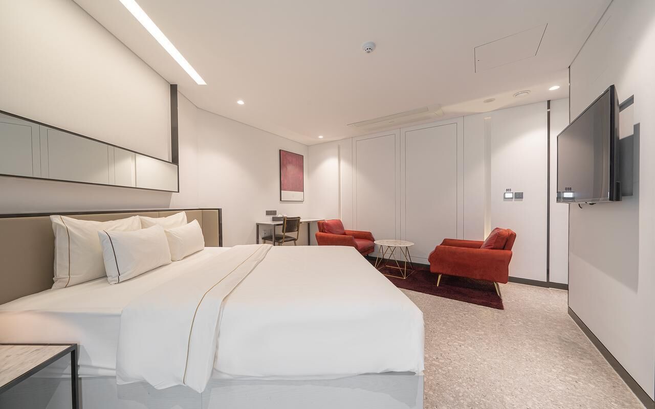 Una habitación de hotel moderna con una cama grande, un televisor montado en la pared y dos sillones rojos.