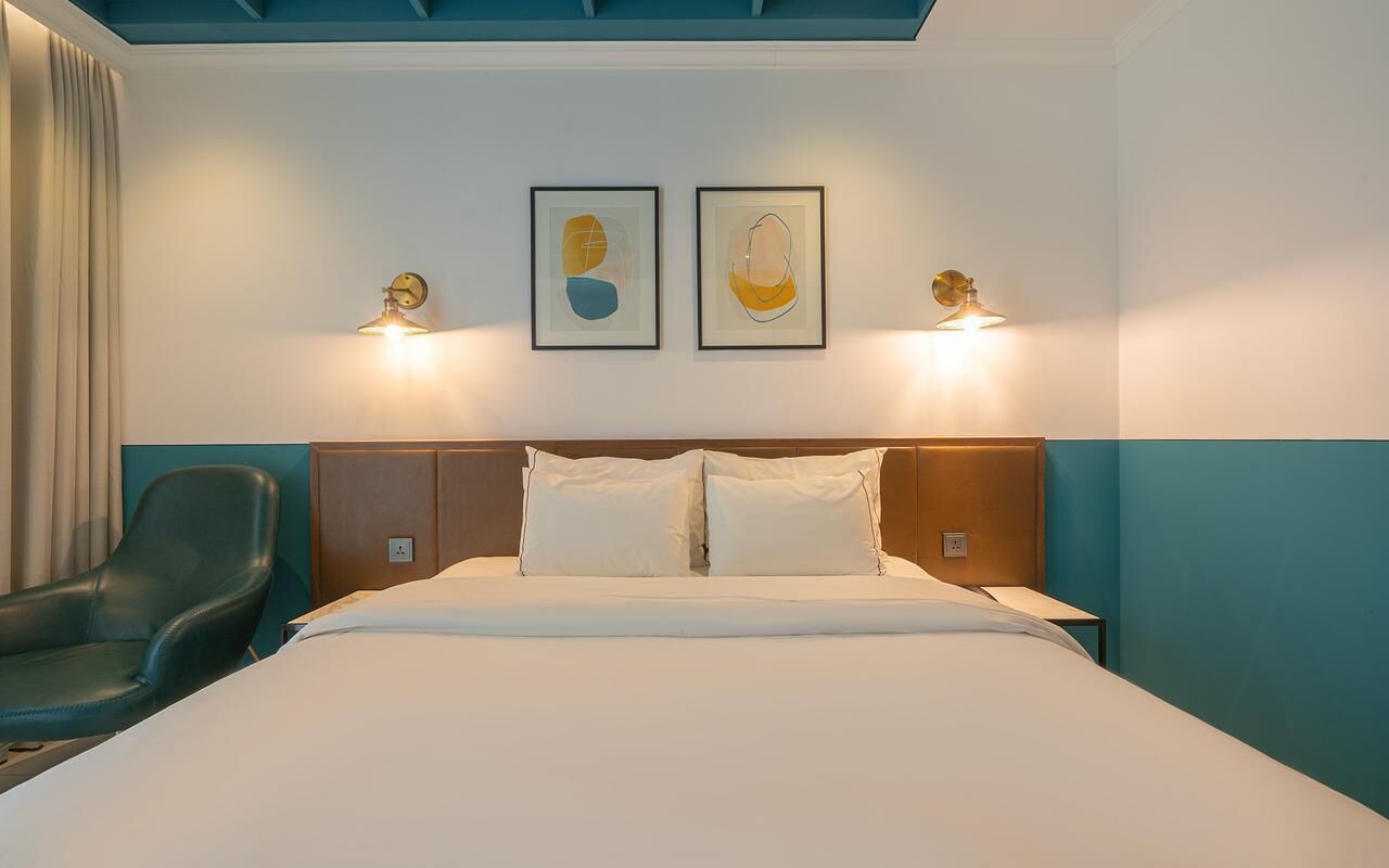 A hotel room with a bed, two wall-mounted lamps, a chair, and two framed artworks.