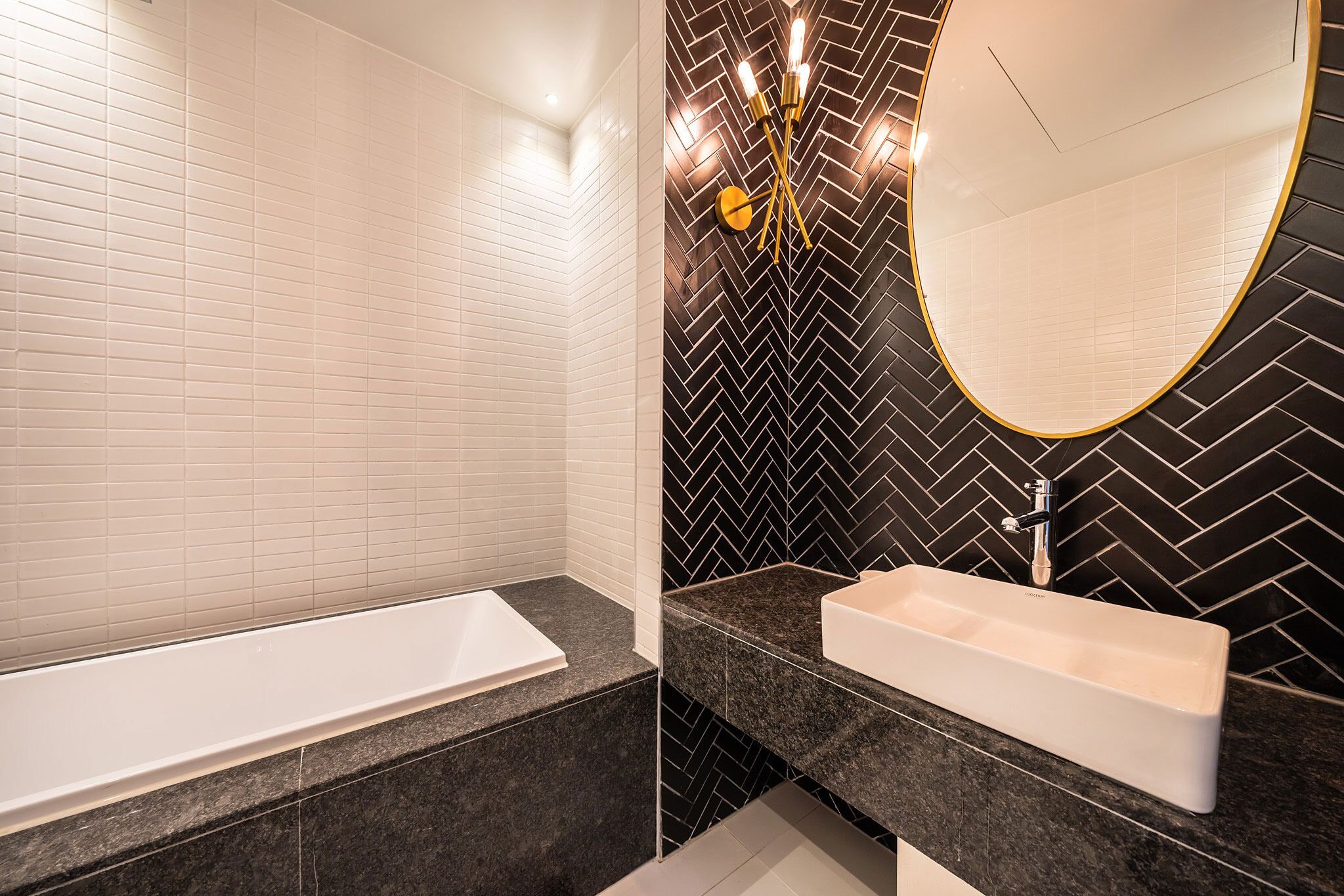 A modern bathroom with a white bathtub, a black herringbone-patterned wall, and a round mirror with a gold frame.