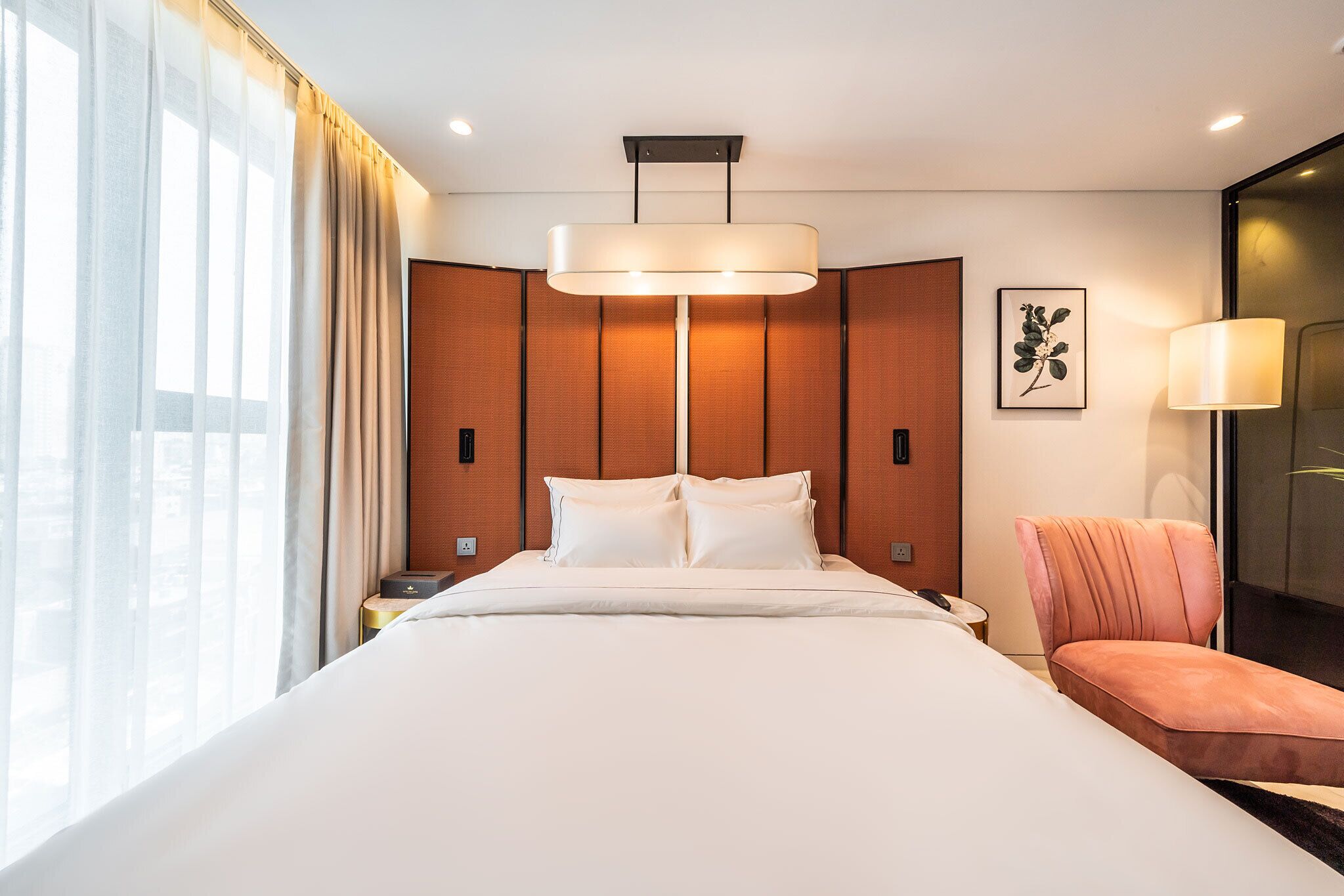 A hotel room with a large bed, a bedside table, a chair, a wall-mounted lamp, and a window with curtains.