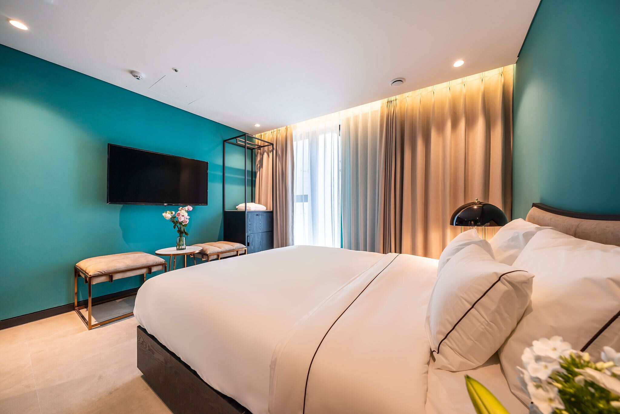 A modern hotel room with a large bed, a flat-screen TV mounted on the wall, a bedside table with a vase of flowers, and a lamp.