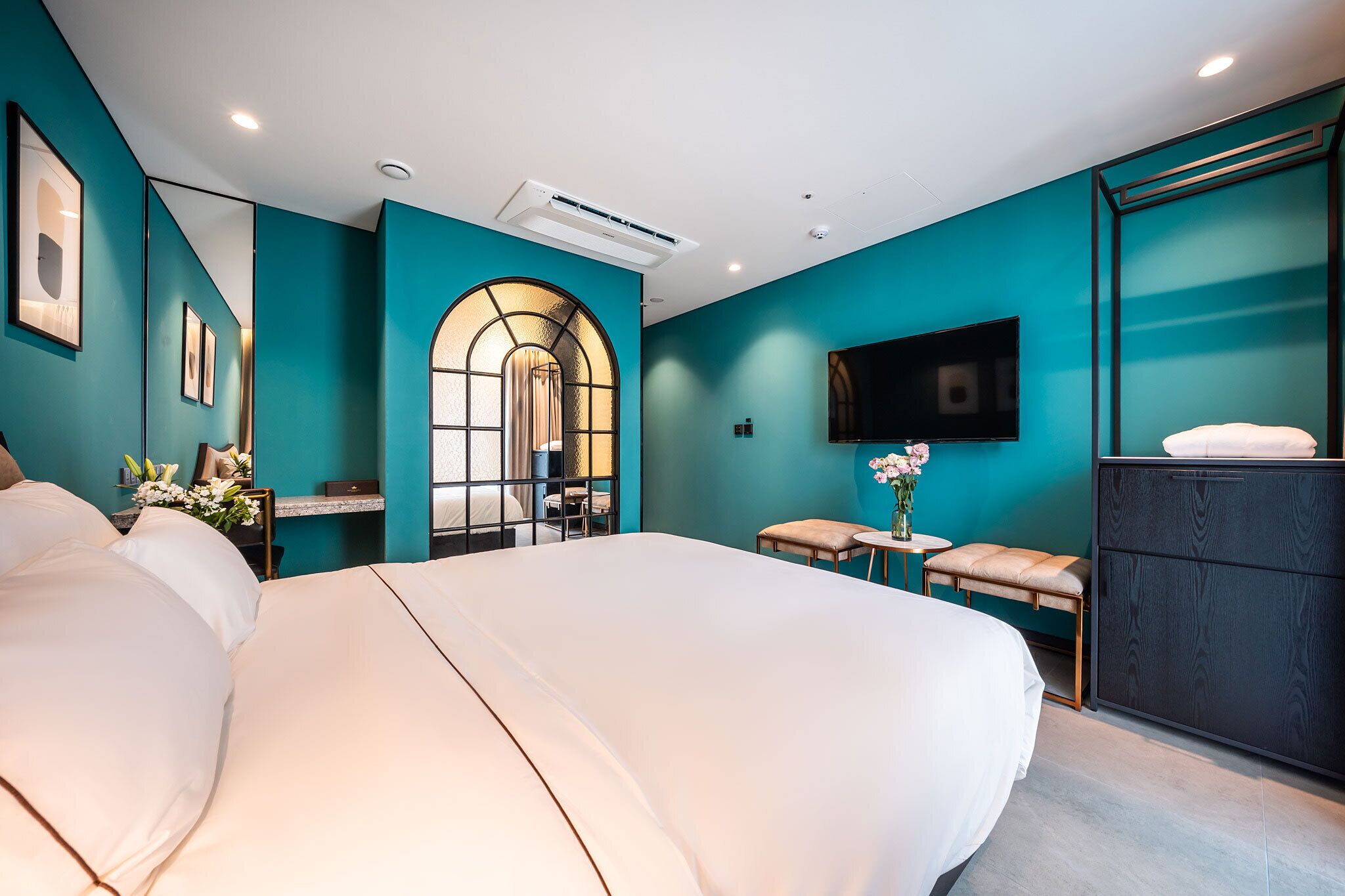 Una habitación de hotel moderna con una cama grande, un escritorio con una silla, una mesita con un jarrón de flores, un banco y un televisor montado en la pared.