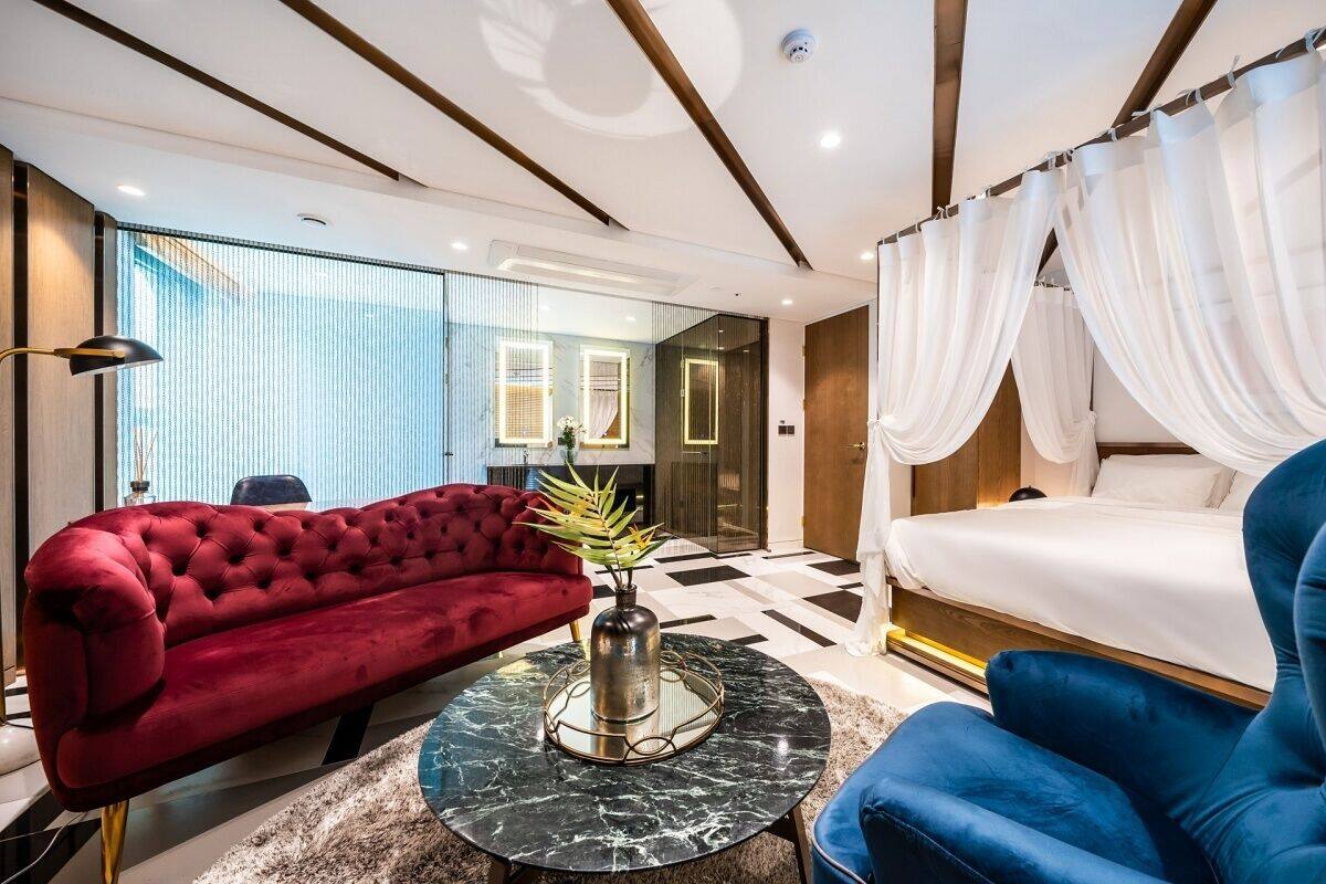 A modern hotel room with a red sofa, a blue armchair, a bed with white curtains, and a marble coffee table.