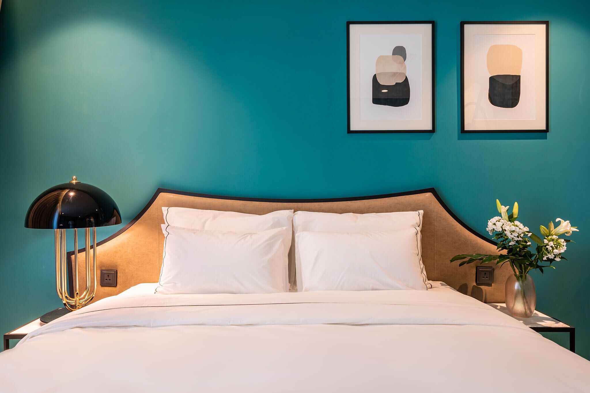 A neatly made bed with white linens, a black lamp with a gold base, and a vase with flowers on a nightstand.