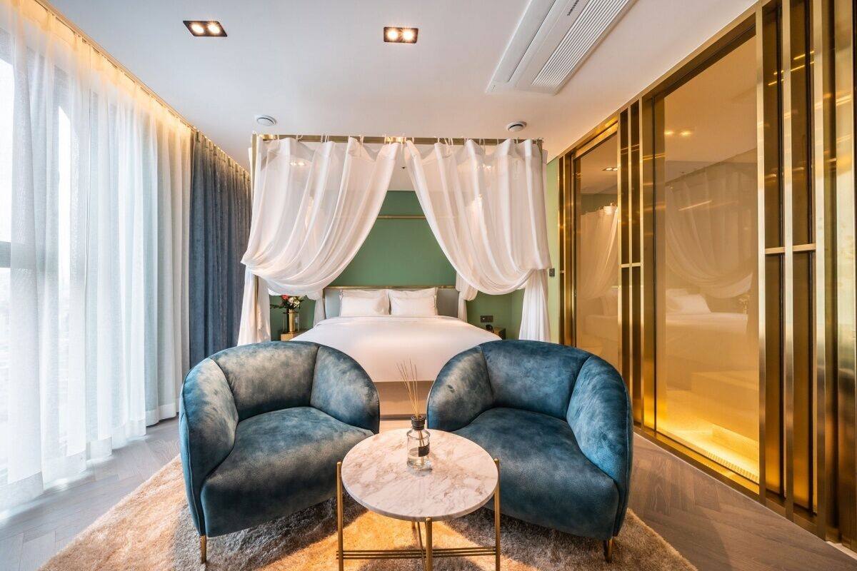 A modern bedroom with a large bed, two armchairs, a small table, and a glass partition.