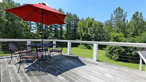 A wooden deck with a table and chairs, a red umbrella, and a view of a grassy area and trees.