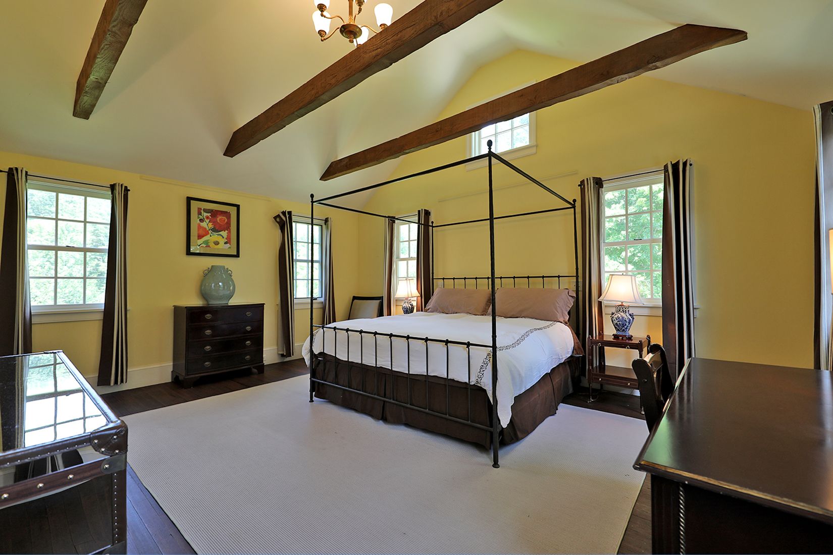 A four-poster bed with a white bedspread, a wooden dresser, a desk, and a window with curtains.