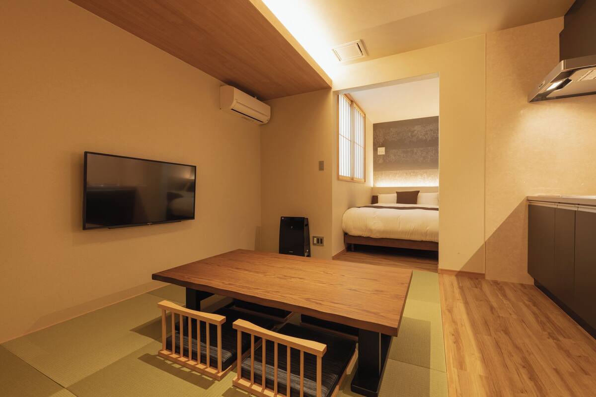 A compact hotel room with a wooden dining table, a TV mounted on the wall, a bed with a window, and a kitchenette area.
