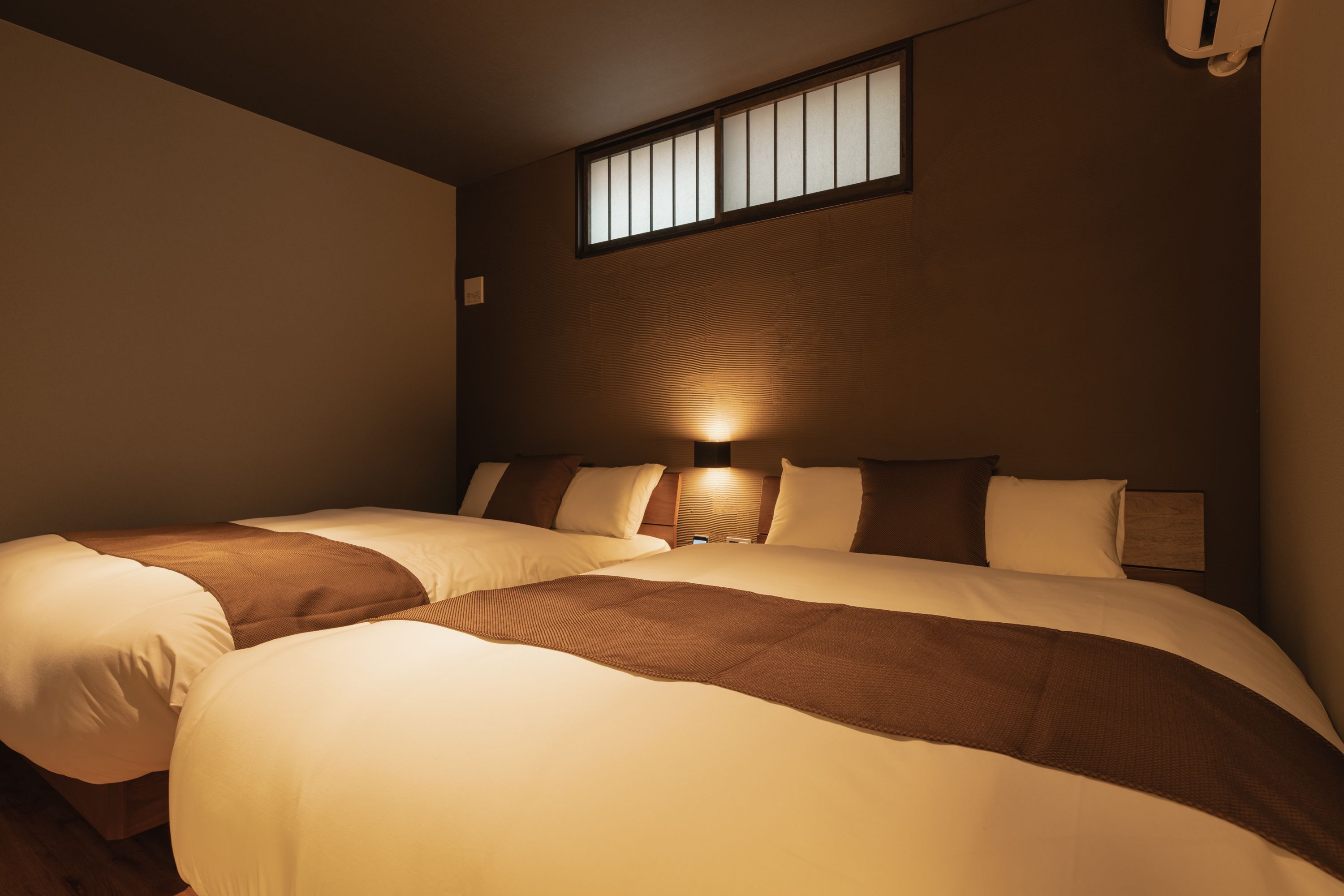 A hotel room with two beds, a window, and a wall-mounted light fixture.