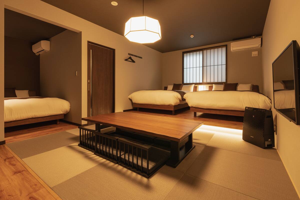 A modern hotel room with a wooden floor, two beds, a large wooden table, and a ceiling light.