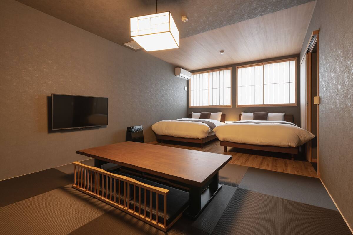A modern hotel room with a flat-screen TV, a wooden table, two beds with pillows, a large window, and a ceiling light fixture.
