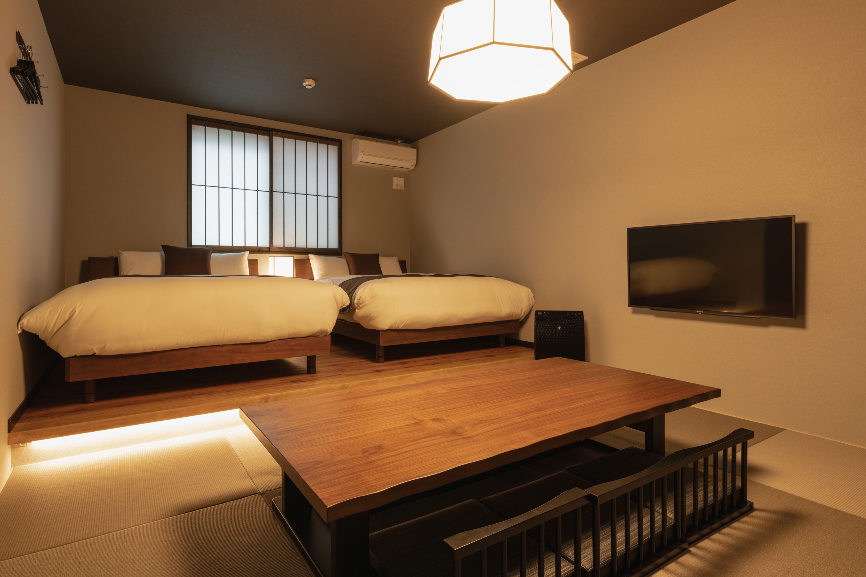 A bedroom with two beds, a wooden table, a TV, and a window with blinds.