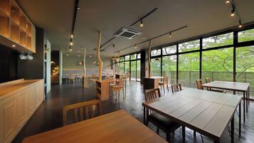 A spacious indoor area with multiple wooden tables and chairs, large windows offering a view of greenery outside, and pendant lighting.