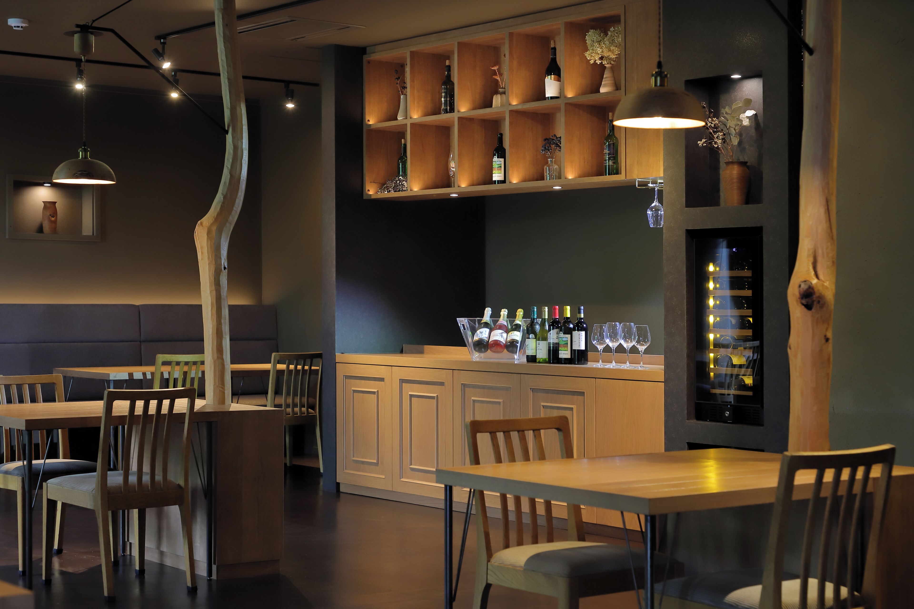A restaurant interior with wooden tables and chairs, a bar area with bottles and glasses, and pendant lighting.