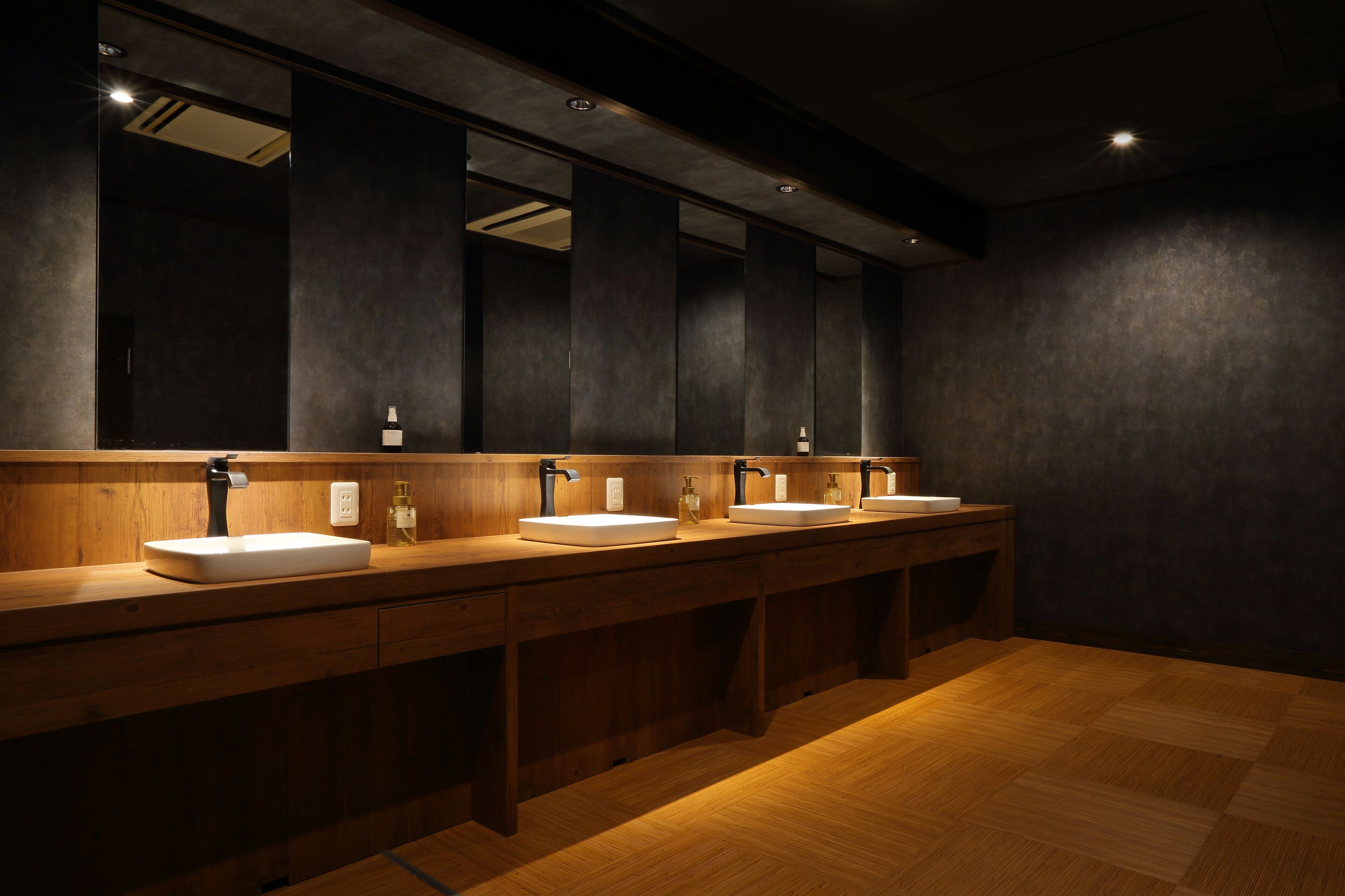 Des toilettes publiques équipées de trois lavabos, de plans-vasques en bois et d’un grand miroir au-dessus de chaque lavabo.