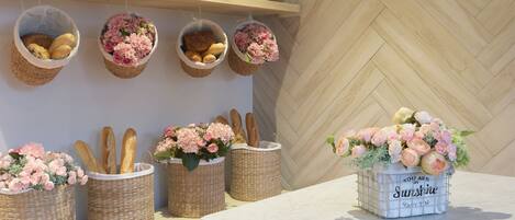 A kitchen with wooden shelves holding baskets of bread and flowers, a marble table, and a herringbone-patterned wall.