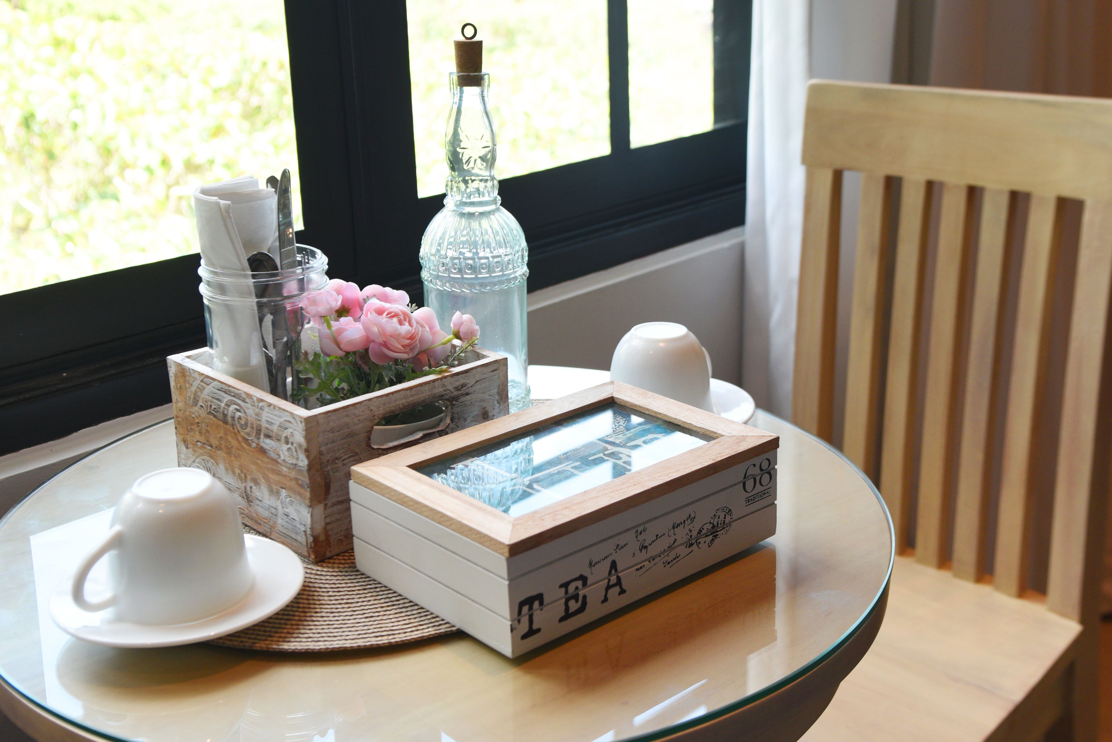 A table with a glass top, a framed box labeled "TEA", a bottle, a container with utensils, and a vase with pink flowers.