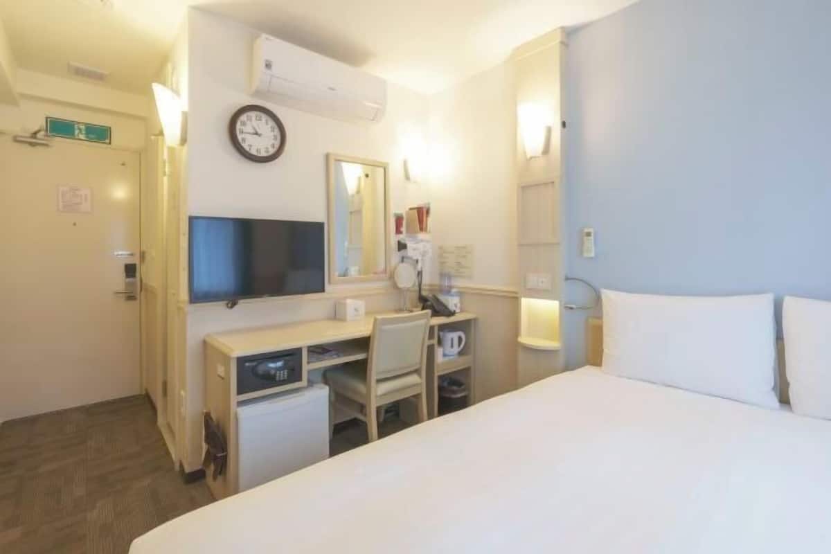 A hotel room with a bed, a desk with a chair, a television, a clock, and a mirror.