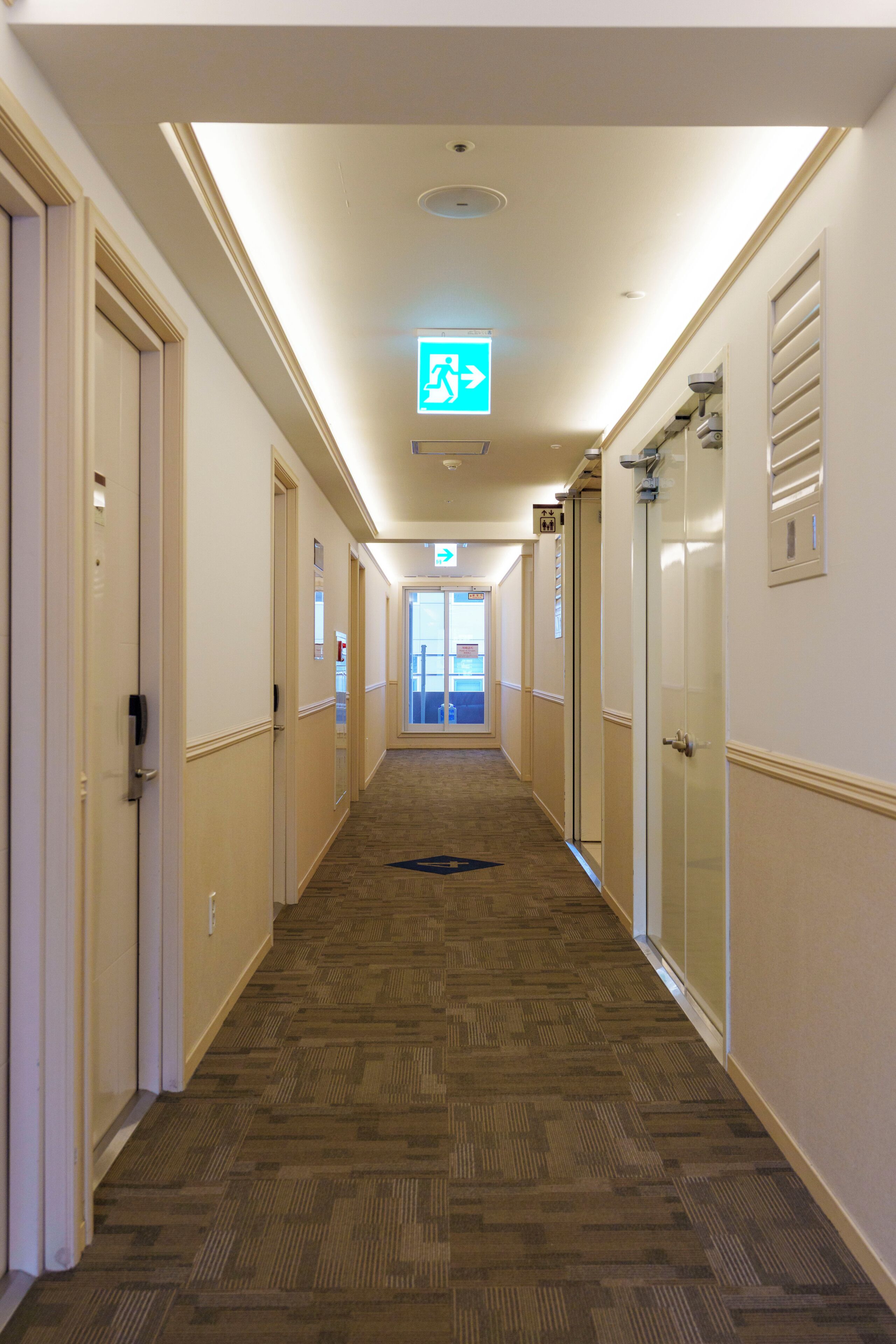 A hotel hallway with a carpeted floor, doors on both sides, and an exit sign on the ceiling.