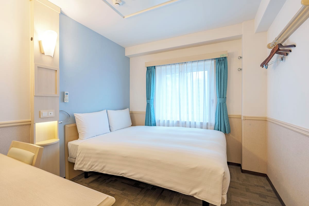 A hotel room with a bed, a desk, a chair, a window with curtains, and a wall-mounted light fixture.