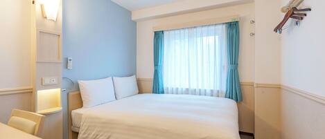 Habitación de hotel con cama, escritorio, silla, ventana con cortinas y lámpara fijada a la pared.