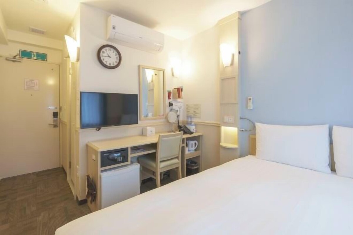 A hotel room with a bed, a desk with a chair, a television, a clock, and a mirror.