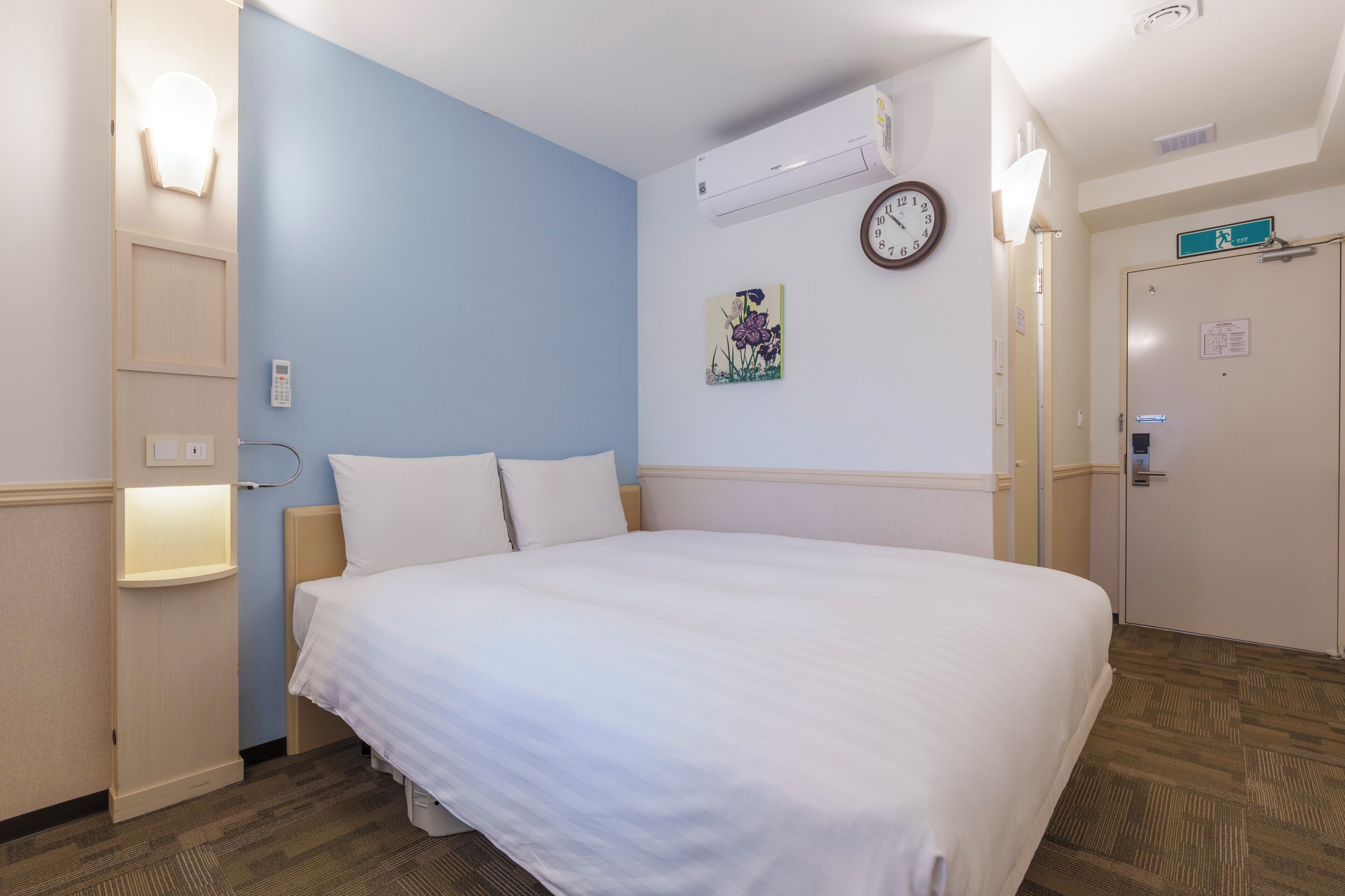 A hotel room with a bed, bedside lamp, wall-mounted air conditioner, clock, and a door with a sign.