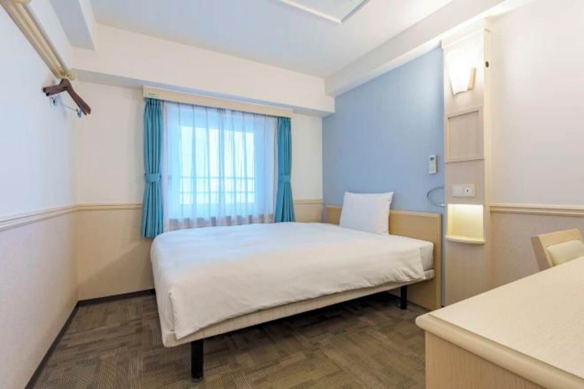 A hotel room with a bed, a bedside table, a chair, a window with curtains, and a wall-mounted shelf.