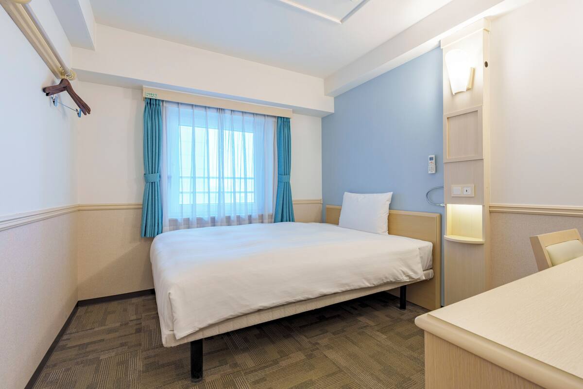 A hotel room with a bed, a bedside table, a chair, a window with curtains, and a wall-mounted light.