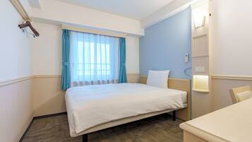 A hotel room with a bed, a bedside table, a chair, a window with curtains, and a wall-mounted light.