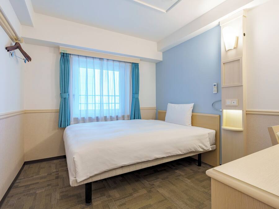Habitación de hotel con cama, mesita de noche, una silla, ventana con cortinas y una lámpara fijada a la pared.