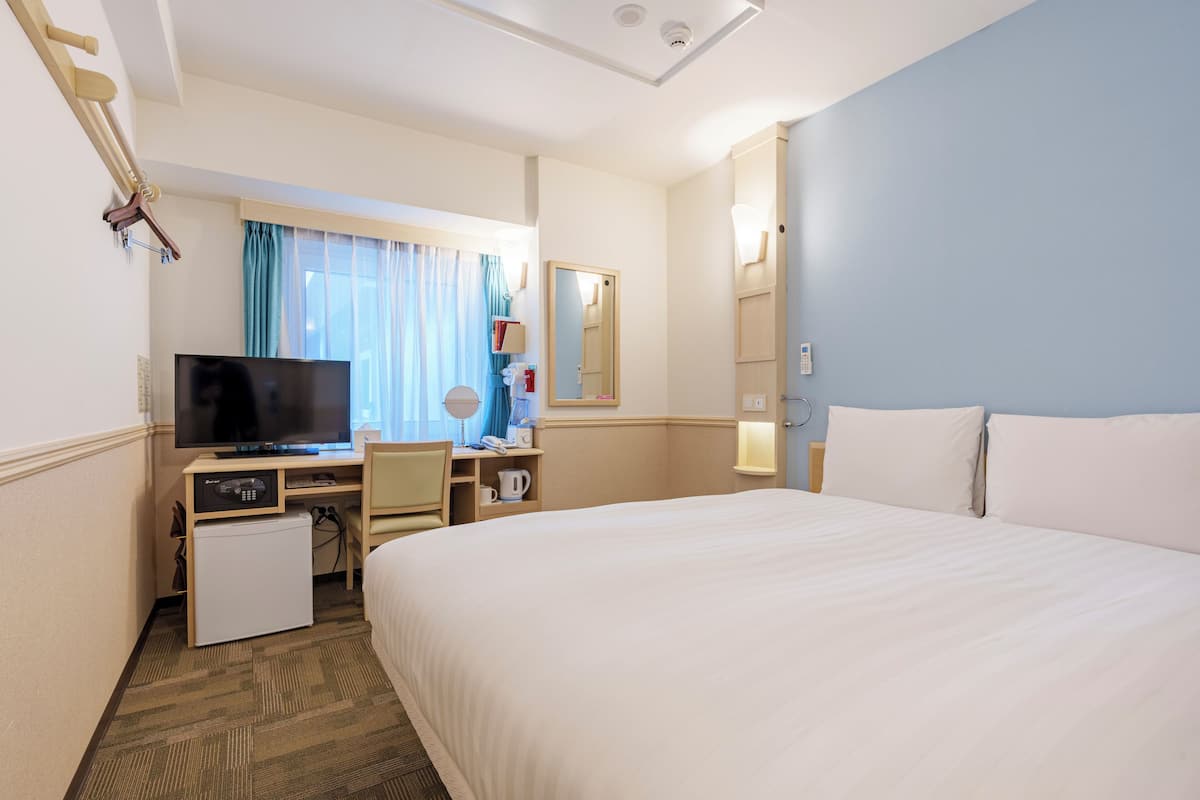 A hotel room with a bed, a desk with a television, a small refrigerator, a mirror, and a window with curtains.