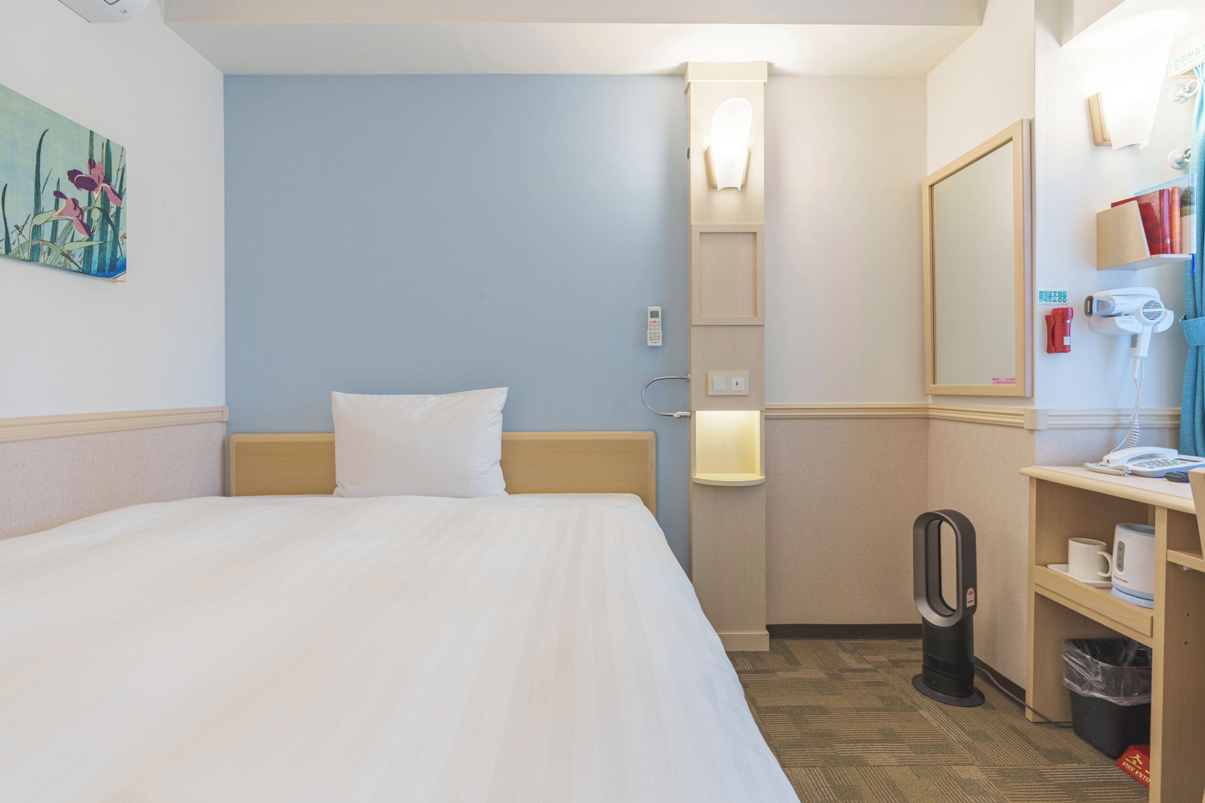 A hotel room with a bed, a bedside table with a lamp, a mirror, a hairdryer, and a trash bin.