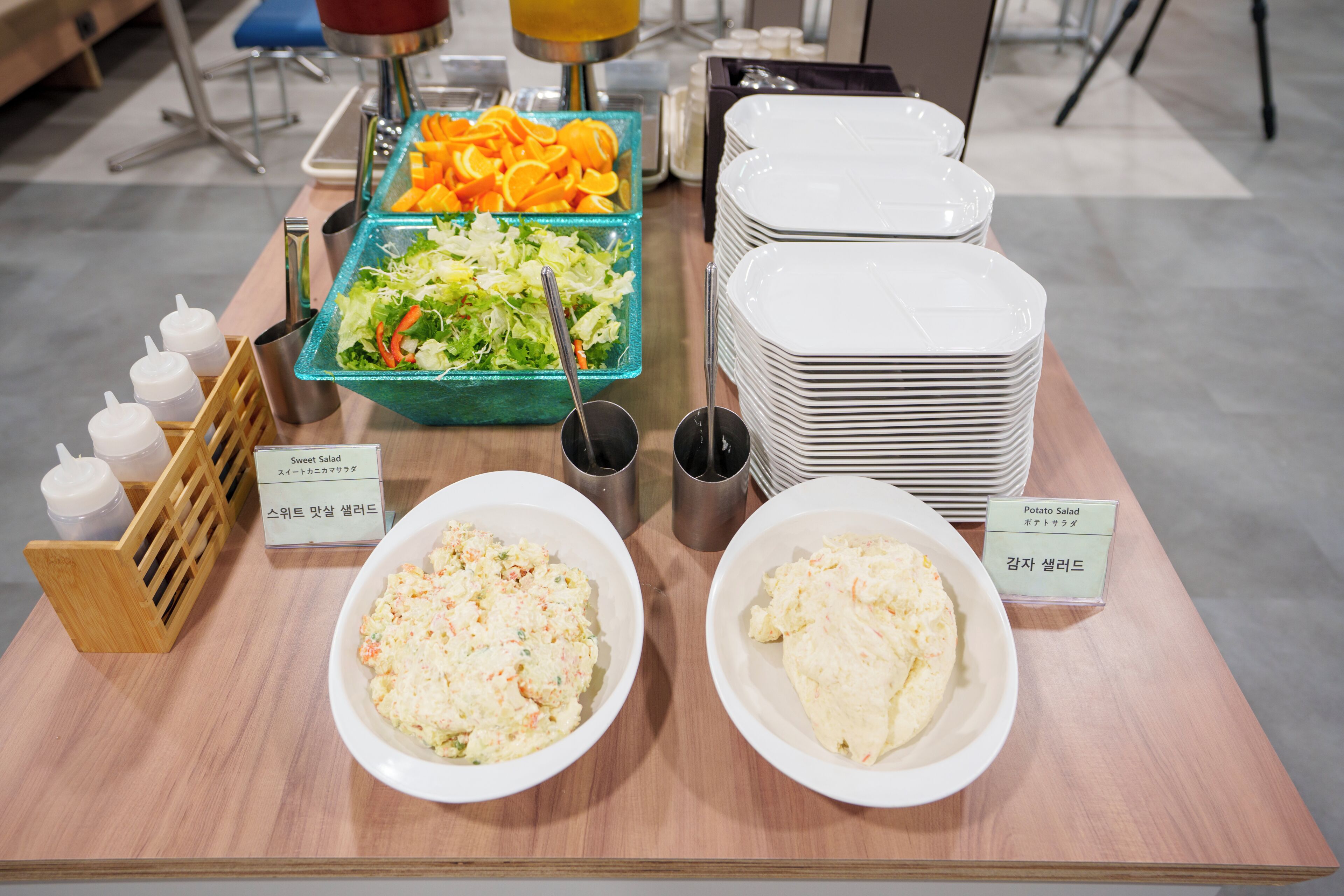 A buffet table with various dishes, including salads and a sweet salad, and a stack of white plates.