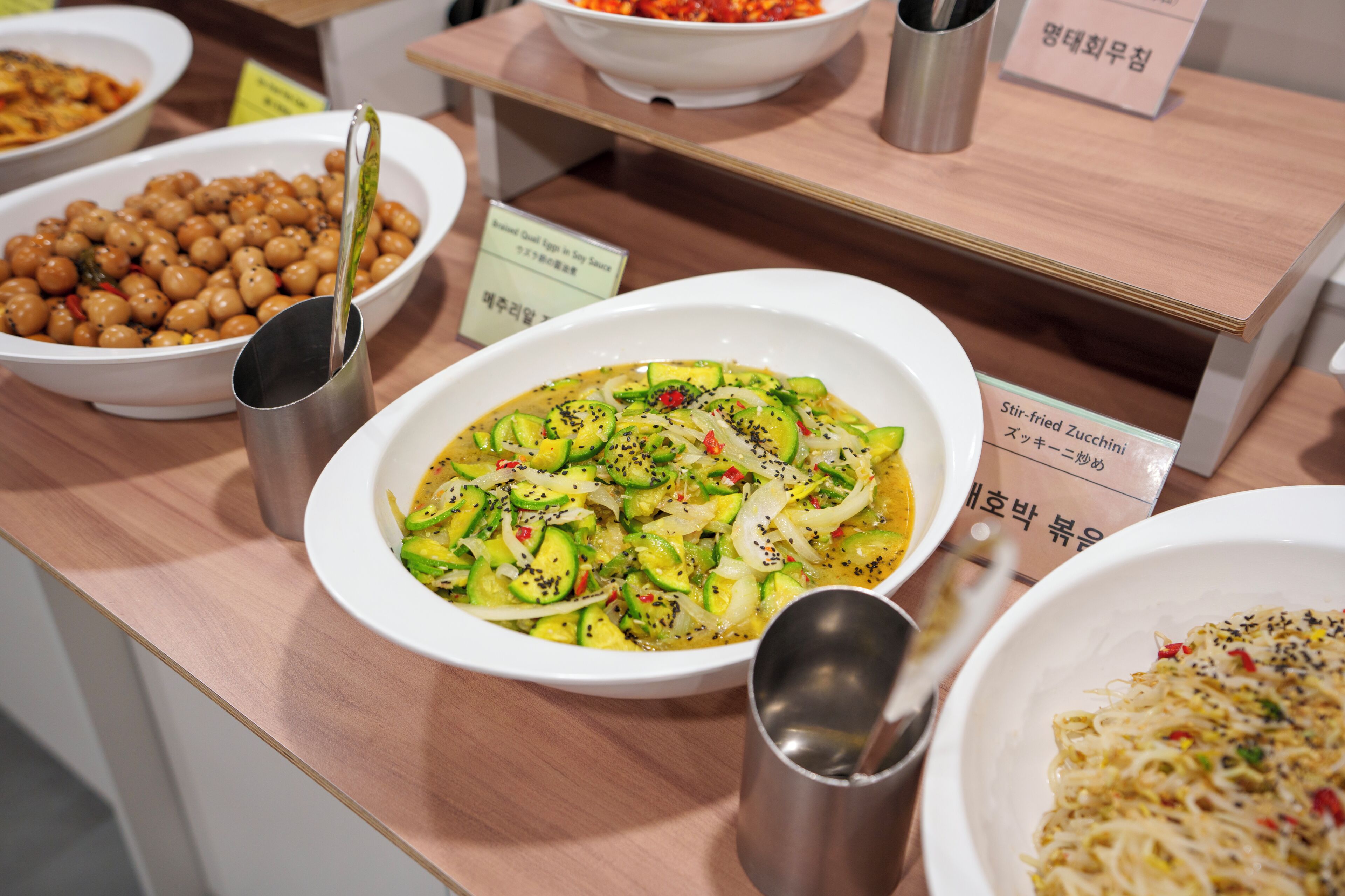 A food display with various dishes, including stir-fried zucchini and chickpeas, and a sign indicating the dish names.