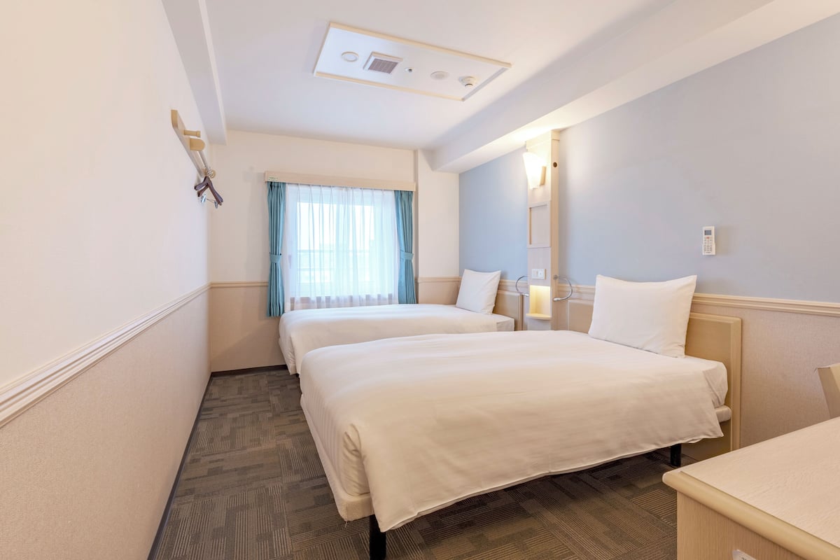 A hotel room with two beds, a window with curtains, a bedside table, a nightstand, and a wall-mounted light.