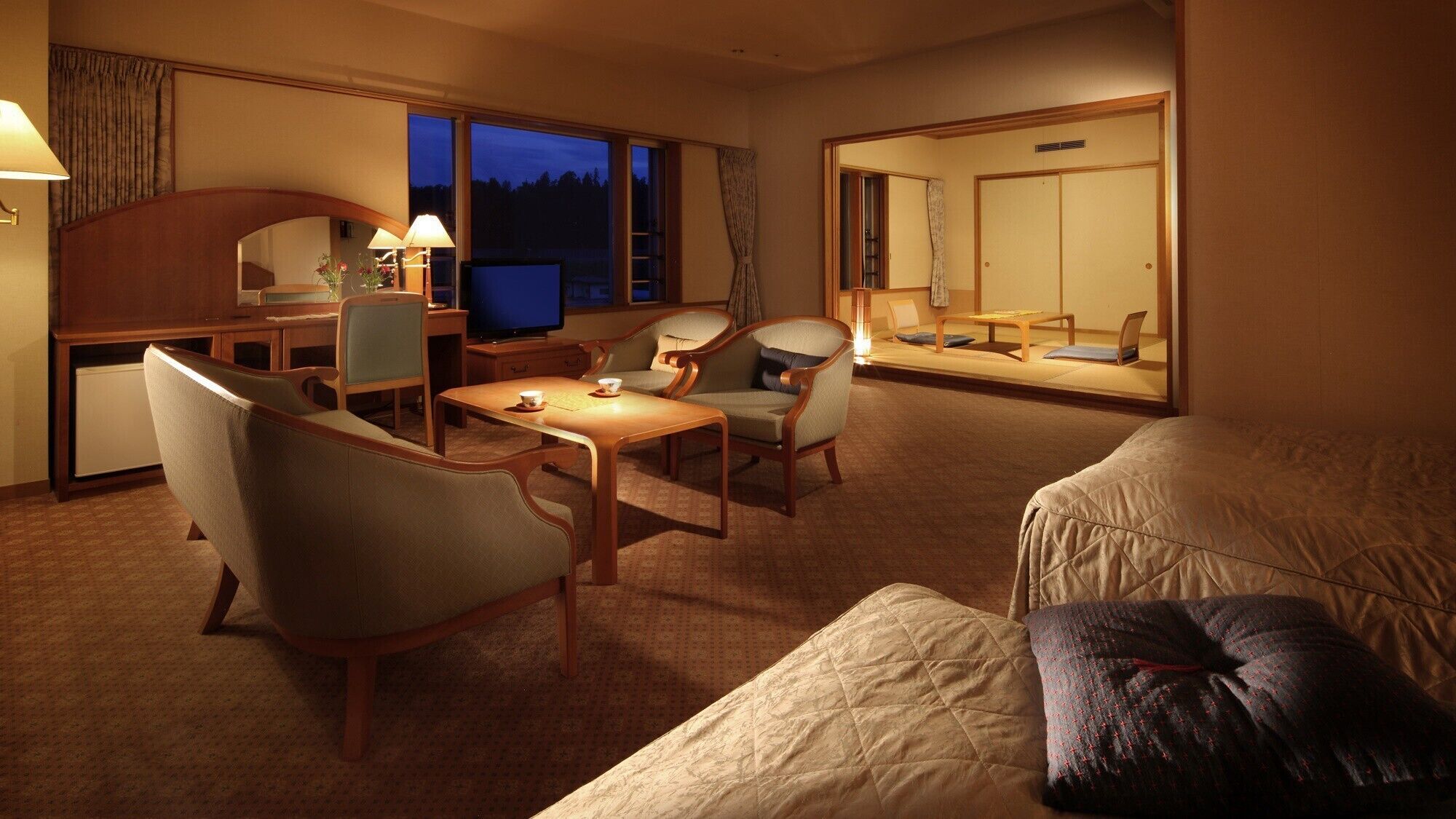 Habitación de hotel con una cama, dos sillones, una mesita, un escritorio con una computadora, un televisor y un espejo grande.