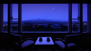 Une pièce avec une table et deux chaises, disposées face à une grande fenêtre offrant une vue sur les montagnes et un croissant de lune.