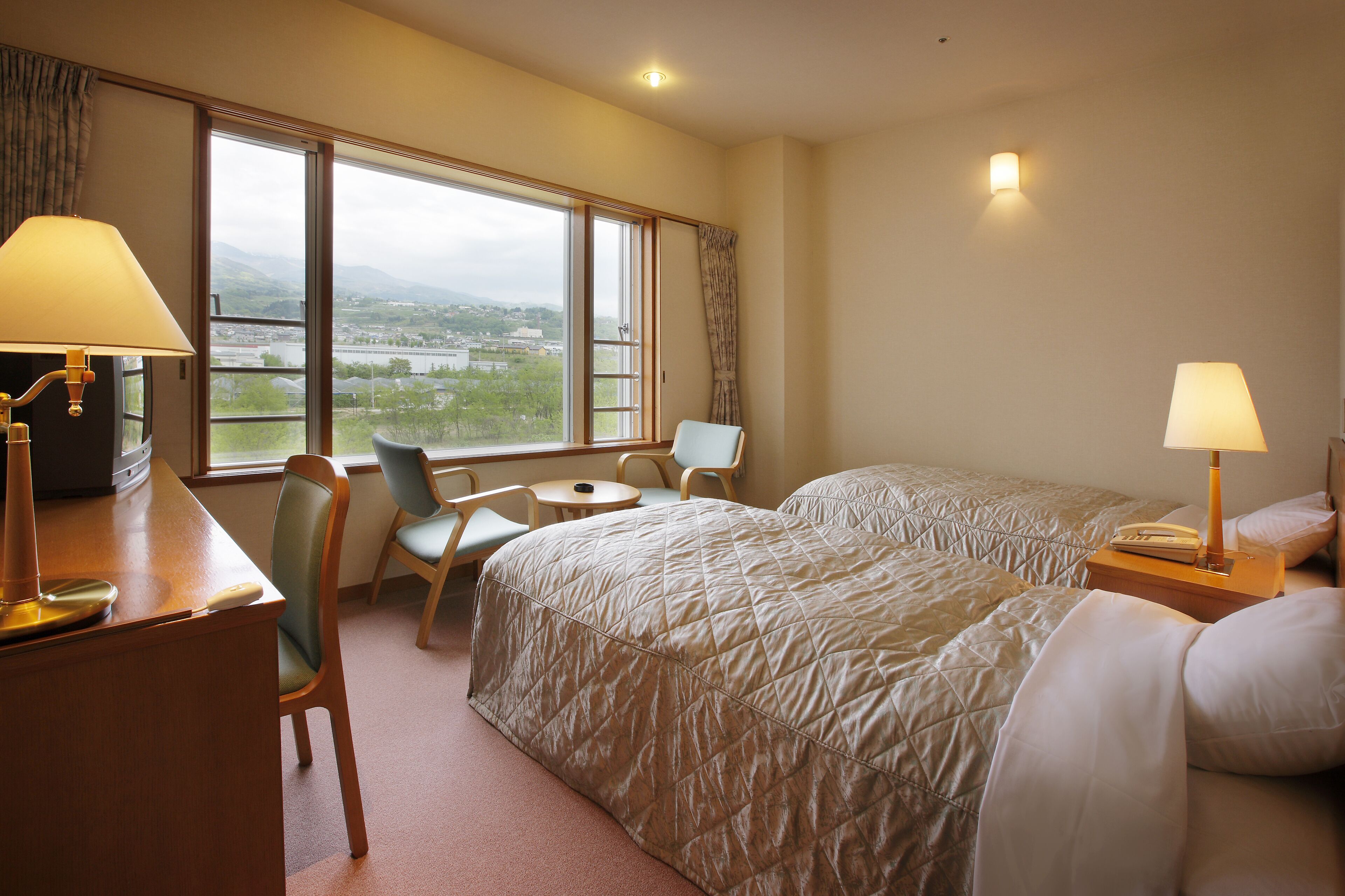 A hotel room with two beds, a desk with a lamp, a chair, a small table, and a view of the outdoors.