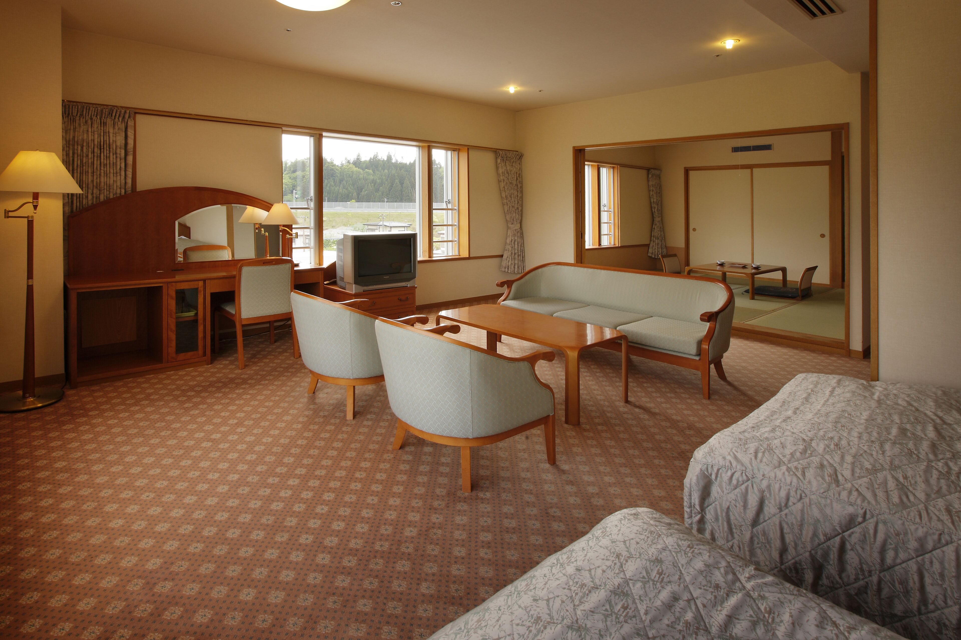 A hotel room with a sofa, armchair, dining table, TV, and a desk with a lamp.