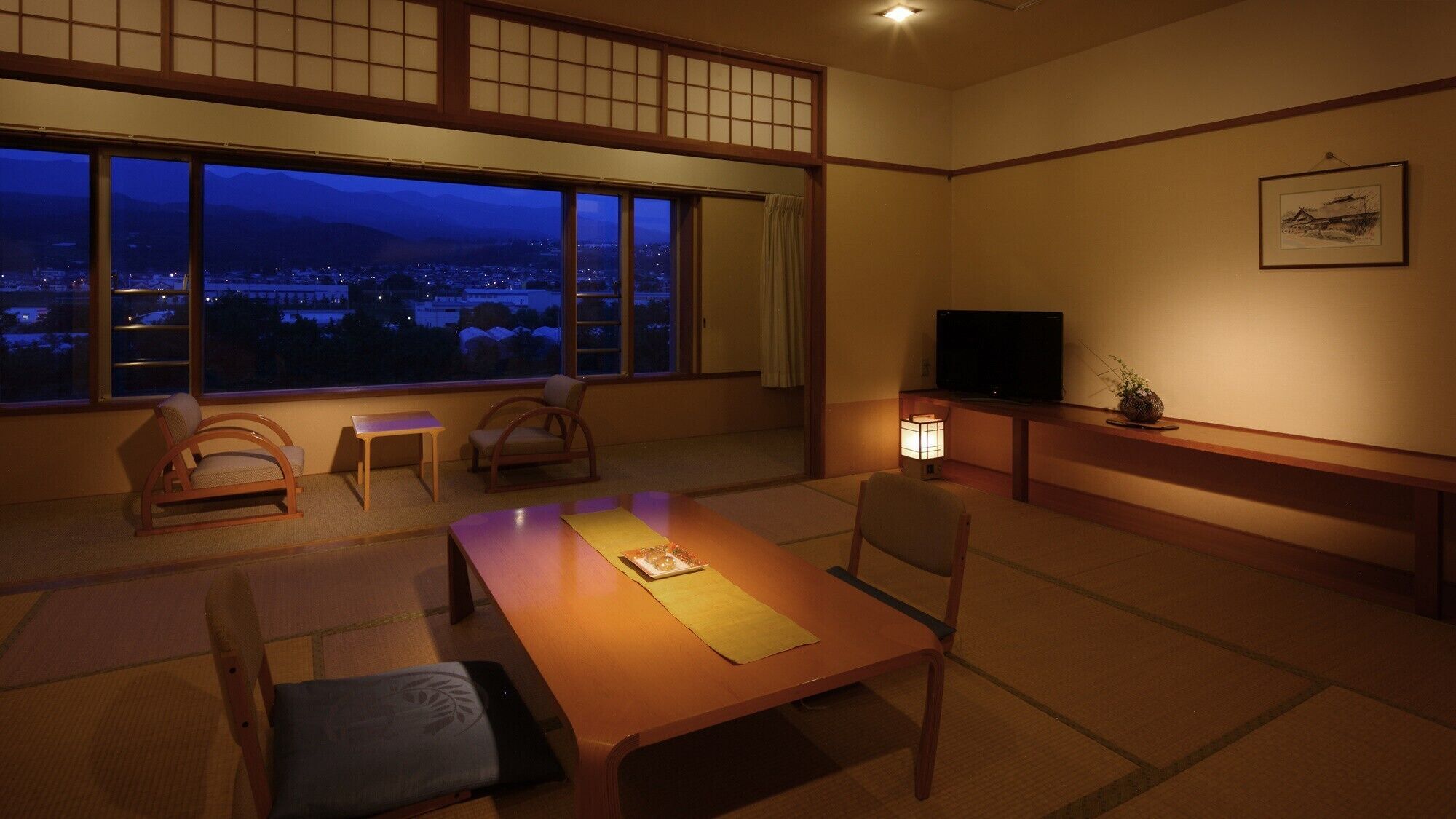 Une pièce de style japonais traditionnel, avec un sol recouvert de tatamis, une table basse, des chaises, un téléviseur et une vue sur le paysage urbain au crépuscule.