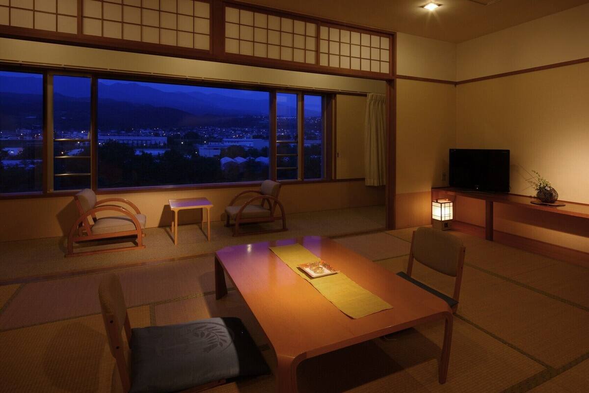 Perinteinen japanilainen huone, jossa on tatamimatto, matala pöytä, tuoleja, televisio ja näkymä kaupunkimaisemaan auringonlaskun aikaan.