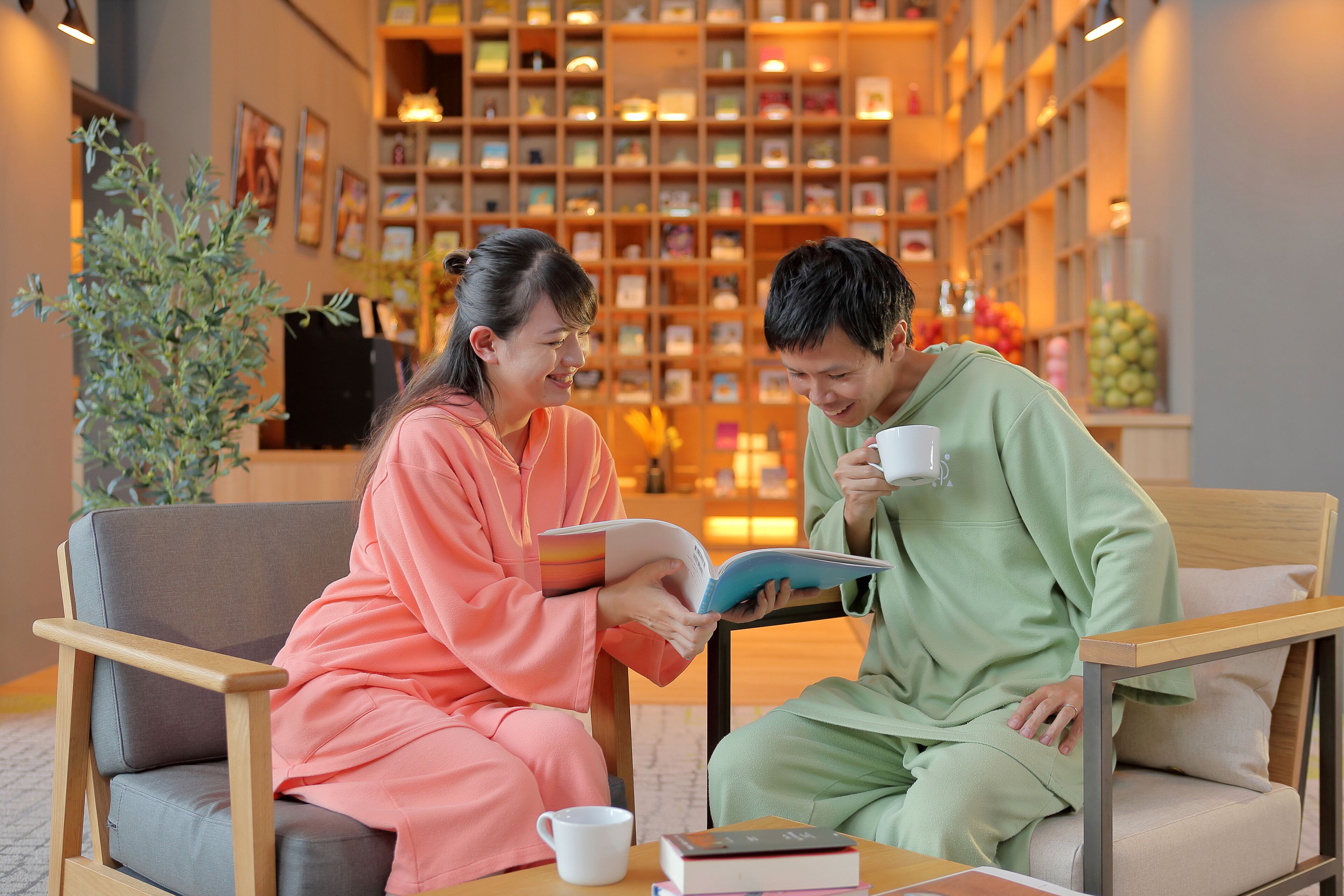 Two people seated on a sofa in a cozy, well-lit room with books and a coffee cup on a table.