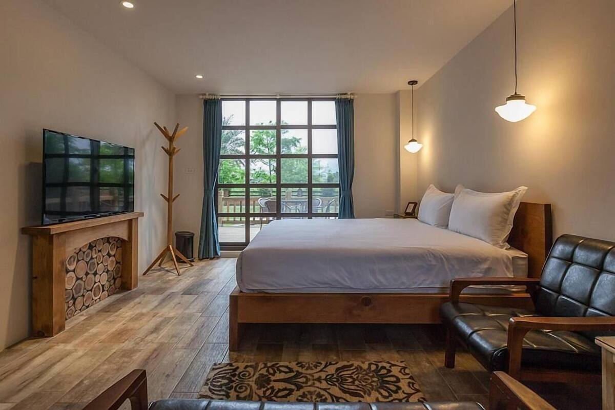 A bedroom with a large bed, a TV on a wooden stand, a window with curtains, and a view of greenery outside.