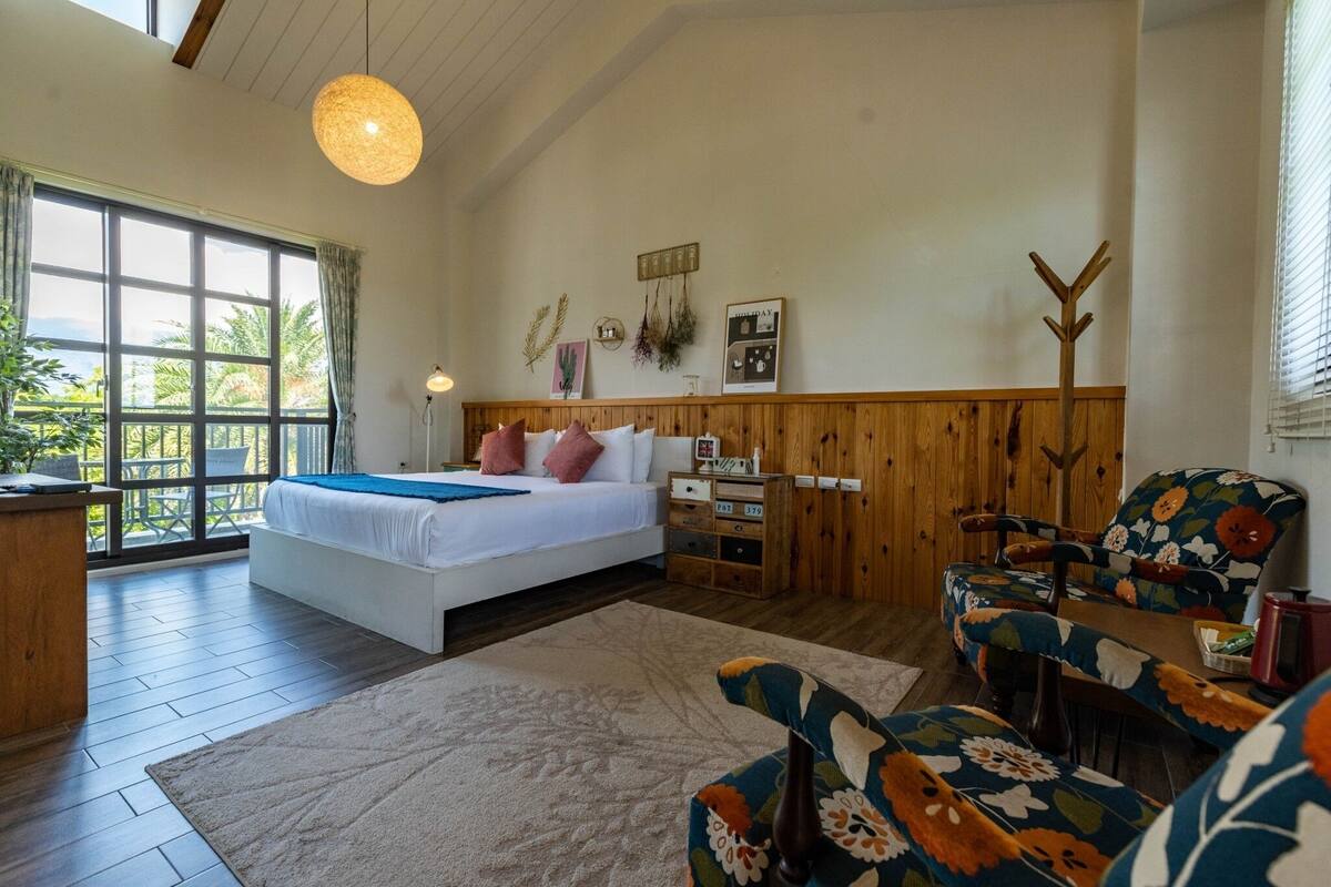 A bedroom with a large bed, wooden headboard, a wooden coat rack, a desk, and a chair with floral upholstery.