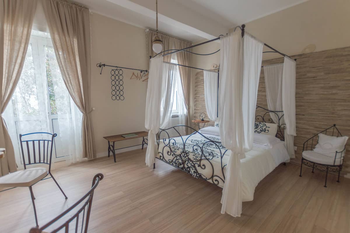 A bedroom with a four-poster bed, a chair, a small table, and a window with curtains.