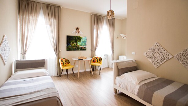 A hotel room with two single beds, a small round table, two chairs, a radiator, and a painting on the wall. - Leucos B&B (Potenza)