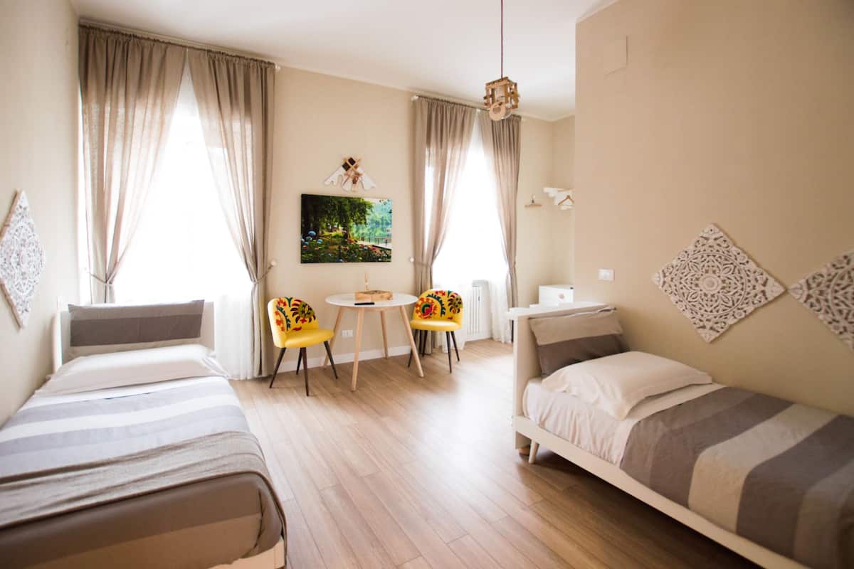 A hotel room with two single beds, a small round table, two chairs, a radiator, and a painting on the wall.